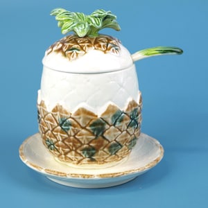 May include: A ceramic pineapple-shaped sugar bowl with a matching lid and spoon, resting on a small saucer. The bowl is white with brown and green accents, and the lid features green leaves. The set is designed to resemble a pineapple.