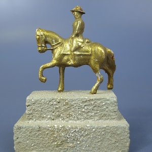 Puede incluir: Una figura de metal de color dorado de un jinete a caballo, montada sobre una base rectangular de color gris claro con textura similar a la piedra. El jinete lleva un sombrero y está sentado en una silla de montar, con el caballo al trote.