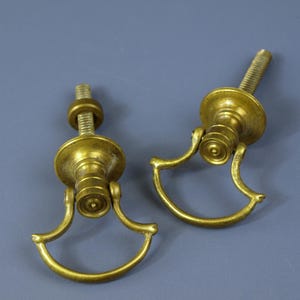 Può includere: Due maniglie antiche per cassetti in ottone con un design decorativo. Ogni maniglia presenta un perno filettato, una base rotonda e un'impugnatura curva. L'ottone ha una calda patina invecchiata, che suggerisce ferramenta vintage o antica.