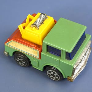 May include: A colorful toy tow truck with a green cab, red bed, and yellow crane. The truck has black wheels and a silver-colored winch. The front bumper and grill are silver. The toy is on a blue background.