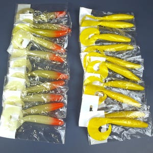 Peut inclure: Deux ensembles d'appâts de pêche dans un emballage plastique transparent. Un ensemble comprend des leurres avec un corps jaune-vert et des pointes orange. L'autre ensemble contient des leurres jaune vif avec une queue bouclée. Les leurres sont disposés sur un fond bleu foncé.