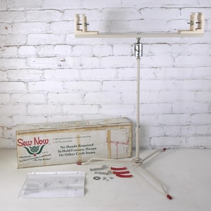 May include: A beige metal craft stand with a tripod base, adjustable height pole, and a horizontal top bar featuring two adjustable wooden clamps. Includes a cardboard box labeled Sew Now, a set of small metal hardware, and a white instruction sheet.