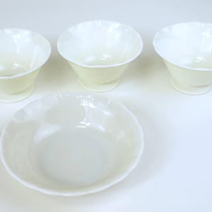 Puede incluir: Cuatro cuencos de color blanco roto con bordes festoneados. Tres son cuencos pequeños con pie y uno es un cuenco más grande y poco profundo. Los cuencos tienen un acabado brillante y están dispuestos sobre una superficie blanca. Para servir.