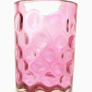 May include: A clear, pink glass tumbler with a textured, bubble-like pattern. The glass has a wavy base and a smooth rim. The color is a vibrant, translucent pink, suitable for beverages.