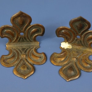 May include: Two antique bronze-colored metal decorative hardware pieces. Each piece has a fleur-de-lis design with a central hole for mounting. One piece has a small area of missing finish.