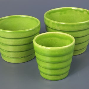 May include: Three lime green ceramic pots with a glossy finish and a distinct ribbed, tiered design. Each pot features a wide circular opening at the top and a tapered base, showcasing a consistent bright green glaze across the exterior surfaces.