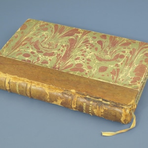 May include: Antique book with a marbled cover in shades of green and red. The spine is brown with visible text and a yellow ribbon bookmark. The book's edges show signs of wear, suggesting its age.