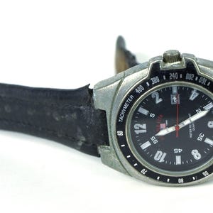 May include: A black-faced wristwatch with a silver metal band and black leather strap. The watch face has white markings and a red second hand. The watch face also has the words "TACHYMETER" and "WATER RESISTANT" printed on it.