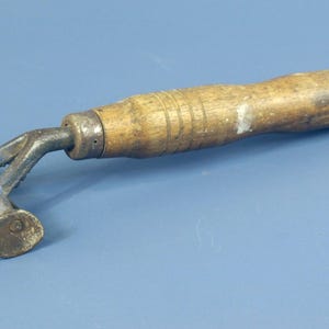 Puede incluir: Una herramienta manual antigua con un mango cilíndrico de madera que presenta ranuras horizontales y un acabado desgastado. La cabeza está hecha de metal oscuro oxidado con un marco en forma de T y un pequeño rodillo cilíndrico giratorio.