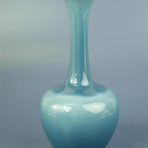May include: A tall, turquoise-colored ceramic vase with a flared rim and a rounded body. The vase has a smooth, glossy finish and a long, slender neck. The vase is a decorative item.