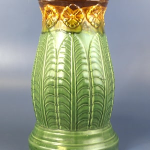 Puede incluir: Un jarrón de cerámica con cuerpo verde brillante y un patrón de hojas verticales en relieve. La sección superior muestra una banda de motivos florales grabados en tono marrón dorado, que termina en un borde esmaltado marrón oscuro.