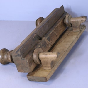 May include: Antique wooden hand plane with two wooden handles and a metal blade. The plane is made of light and dark wood and has a worn finish.