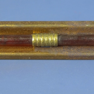 Könnte beinhalten: Eine rechteckige Messingstange mit gebogenem Profil und einem mittigen, horizontalen, dunkelroten zylindrischen Stab. Der Stab wird durch ein kleines, geripptes Messingband in der Mitte gehalten. Die Messingoberfläche weist eine gealterte Patina auf.