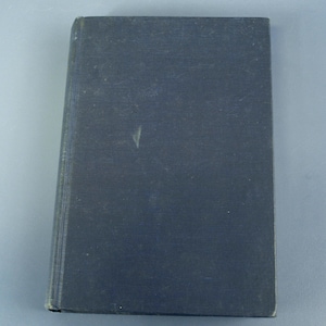 May include: A closed, hardcover book with a dark blue cover. The book's cover has a slightly textured appearance and shows signs of wear. The spine is visible on the left side, indicating the book's pages. The book is set against a light gray background.