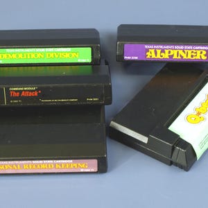 Könnte beinhalten: Eine Sammlung schwarzer Texas Instruments Solid-State-Cartridges. Die Etiketten zeigen Spieletitel wie "Demolition Division", "Alpiner", "The Attack", "Personal Record Keeping" und "Q*bert". Die Cartridges haben Etiketten in verschiedenen Farben.
