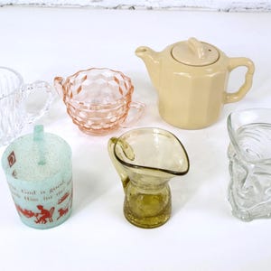Op de afbeelding: Een verzameling van zes vintage glazen kannen en een theepot. De kannen zijn in verschillende vormen en maten, waaronder een heldere glazen kan met een geribbeld ontwerp, een roze glazen kan met een diamantpatroon, een heldere glazen kan met een schedelontwerp, een heldere glazen kan met een textuur en een gele glazen kan met een afgeronde basis. De theepot is crèmekleurig met een deksel en een tuit.