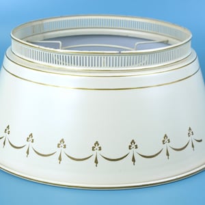May include: A cream-colored metal Christmas tree collar with a tapered, conical shape. The collar features a gold-toned decorative garland pattern around the base and a perforated, fence-like rim at the top with a gold-toned wire frame inside.