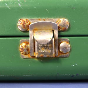 May include: Close-up of a green metal box with a tarnished metal latch. The latch is marked with the text "HARTFORD CONN NIELSEN HARDWARE CORP" and is secured by four rusty rivets.