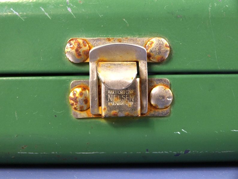 May include: Close-up of a green metal box with a tarnished metal latch. The latch is marked with the text "HARTFORD CONN NIELSEN HARDWARE CORP" and is secured by four rusty rivets.