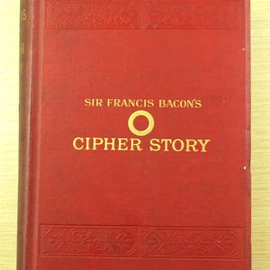 May include: A vintage book with a red cover featuring the title "CIPHER STORY" in gold lettering. The author's name, "SIR FRANCIS BACON'S," is also in gold. Decorative embossed patterns are visible on the cover. The book is resting on a light-colored surface.