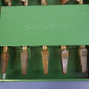 May include: A vintage green metal tool case with the brand name "GREENLEE" printed on the front. The case contains several metal drill bits, each labeled with a number. The case shows signs of wear and tear.