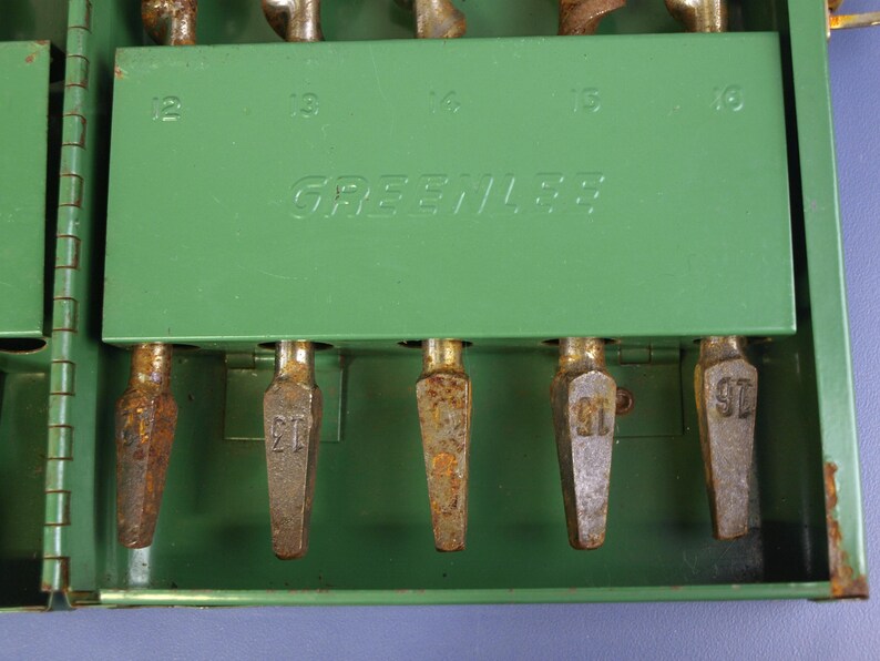 May include: A vintage green metal tool case with the brand name "GREENLEE" printed on the front. The case contains several metal drill bits, each labeled with a number. The case shows signs of wear and tear.