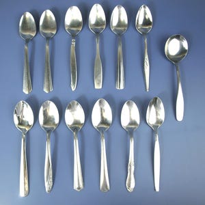 Puede incluir: Una colección de doce cucharas de plata de varios diseños, dispuestas sobre una superficie azul. Las cucharas incluyen diferentes formas de mango y tamaños de cuenco, lo que refleja diversos estilos. Las cucharas están hechas de un metal brillante y reflectante.