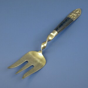 May include: A silver serving fork with a twisted stem and a dark wooden handle. The handle is capped with a silver design. The fork has three tines and is set against a blue background.