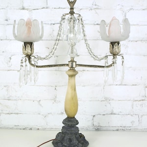 May include: An antique table lamp with a decorative base and a cream-colored column. The lamp features two frosted glass shades with pink interiors, crystal accents, and a crystal finial. The lamp is wired with a brown cord.