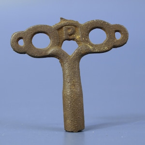 May include: A brown, textured metal winding key featuring a T-shaped handle with two circular cutouts and a central letter P embossed on the top section. The key has a cylindrical shaft and a weathered, oxidized appearance throughout its surface.