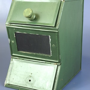 May include: A vintage green metal box with two hinged compartments and a small, round wooden knob. The box has a rectangular black window and a slightly angled design. The paint shows signs of wear, giving it a rustic appearance.