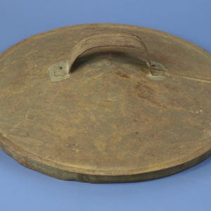 May include: A round, antique metal lid with a handle. The lid is a weathered brown color with a slightly textured surface. The handle is a curved strip of the same metal, attached with rivets.