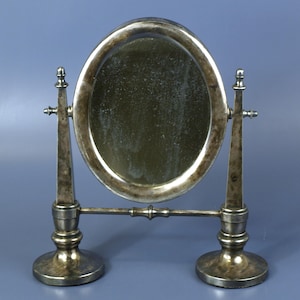 May include: A silver-toned oval tabletop mirror featuring a weathered, tarnished finish. The mirror is supported by two vertical pillars with decorative finials, connected by a horizontal bar, and rests on two circular, weighted pedestal bases.