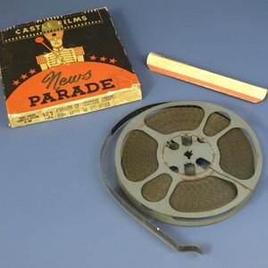 May include: Vintage film reel with a Castle Films "News Parade" box. The box is black with orange lettering and a graphic of a marching band member. A small, rectangular box with red and yellow stripes is also present.