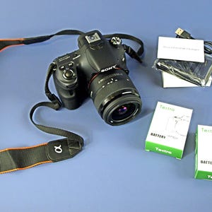 Pode incluir: Câmera digital Sony preta com lente, alça e acessórios, incluindo duas caixas de bateria, um cabo USB e um carregador. A alça da câmera é preta com detalhes em laranja e o logotipo da Sony.