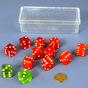 May include: A collection of red and green dice with white dots, alongside a clear plastic box and a penny. The dice are scattered on a blue surface. The red dice have white dots, and the green dice have white dots.