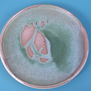 May include: A round ceramic plate featuring a smooth, light pink base color with a textured, mottled green glaze overlay. The green glaze is applied in an irregular, abstract pattern that leaves sections of the pink clay visible.