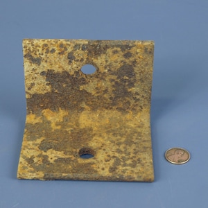 Puede incluir: Un soporte metálico en ángulo recto y oxidado, con dos agujeros, probablemente para montaje. El soporte tiene una superficie desgastada y texturizada con tonos marrones y anaranjados. Se incluye una moneda como referencia de tamaño.