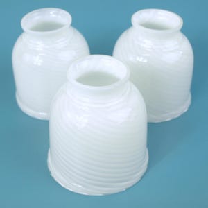 May include: Three white glass lamp shades with a subtle swirling ribbed pattern. Each shade features a rounded, bell-like shape with a circular opening at the top and a wider, open base, showcasing a translucent finish and smooth texture.