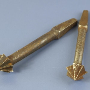 May include: Two metallic countersink drill bits with a weathered, brownish-gold finish. Each tool features a cylindrical shaft, a square-shaped shank at one end, and a multi-fluted conical cutting head at the opposite end for drilling holes.