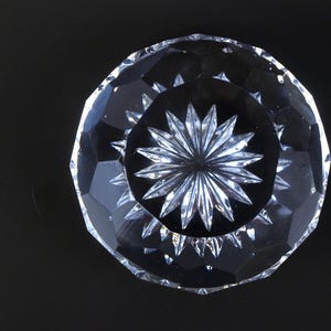 May include: A clear, faceted crystal bowl with a starburst design in the center. The bowl has a circular shape with a series of angled cuts around the rim. The bowl is on a black background.
