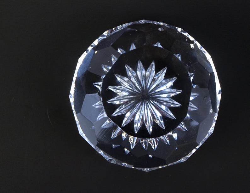 May include: A clear, faceted crystal bowl with a starburst design in the center. The bowl has a circular shape with a series of angled cuts around the rim. The bowl is on a black background.