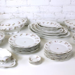 Puede incluir: Un juego completo de vajilla de porcelana blanca con un estampado floral en tonos rosas y verdes. El juego incluye platos, cuencos, fuentes, una salsera y una taza de té con platillo. Los platos están colocados sobre una superficie blanca.