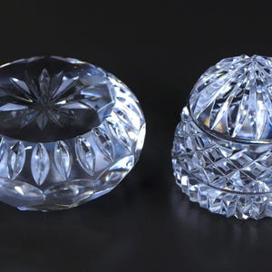 May include: Two clear crystal decorative pieces. One is a round, open bowl with a starburst design in the center. The other is a covered jar with a diamond-cut pattern. Both pieces are on a dark background.