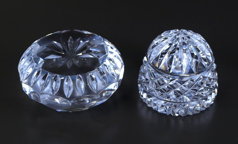 May include: Two clear crystal decorative pieces. One is a round, open bowl with a starburst design in the center. The other is a covered jar with a diamond-cut pattern. Both pieces are on a dark background.