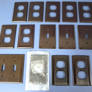 May include: A collection of brown electrical outlet and switch covers. The covers are made of a ridged, metallic material. There are various configurations, including single and double outlet covers, and single switch covers. A package of screws is also included.