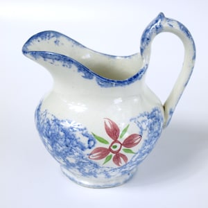 Può includere: Un brocca di ceramica bianca con un motivo a macchie blu e bianche e un disegno di un fiore rosa. La brocca ha un manico e un beccuccio.