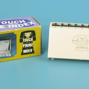 May include: A vintage phone index with a cream-colored plastic case and a box. The box is yellow and purple with the text "ONE TOUCH PHONE INDEX". The index has a retro phone graphic on the front.