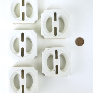 May include: Five white ceramic tiles with a circular design and a central vertical slot, arranged on a white surface. A penny is next to the tiles for size comparison. The tiles have a square shape with rounded corners.