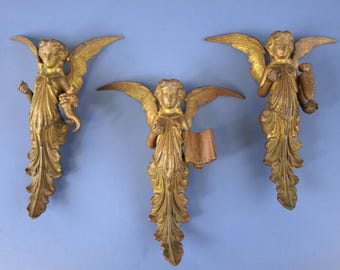 May include: Three gold-toned wall-mounted angel figures with outstretched wings. The left figure holds a cornucopia, the centre figure holds an open book, and the right figure holds a decorative urn. Each figure features flowing robes.